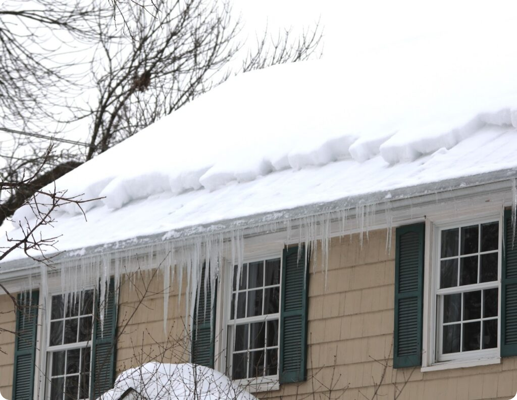 Snow-covered roof with icicles and ice dams forming along the eaves of a home. This photo shows how roof ice buildup occurs without gutter heat tape, emphasizing the importance of self-regulating heat cables like Heat Tape PRO to prevent water damage and keep gutters clear in winter.